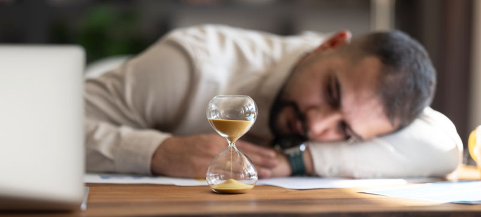 A businessman in a smart shirt sits with his head on his desk, next to his computer, and watches sand falling through an hour glass. A businessman in a smart shirt sits with his head on his desk, next to his computer, and watches sand falling through an hour glass.