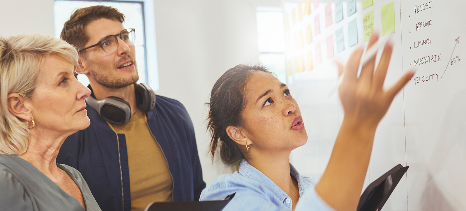 A young woman points to a whiteboard with an older lady and man looking at it A young woman points to a whiteboard with an older lady and man looking at it