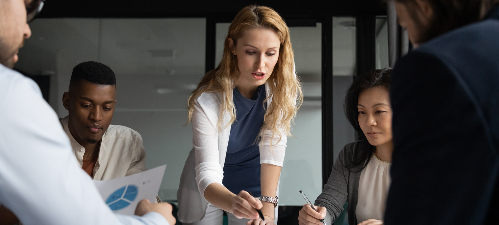 A woman stands up and points to the table with her pen as others in a business meeting look on A woman stands up and points to the table with her pen as others in a business meeting look on