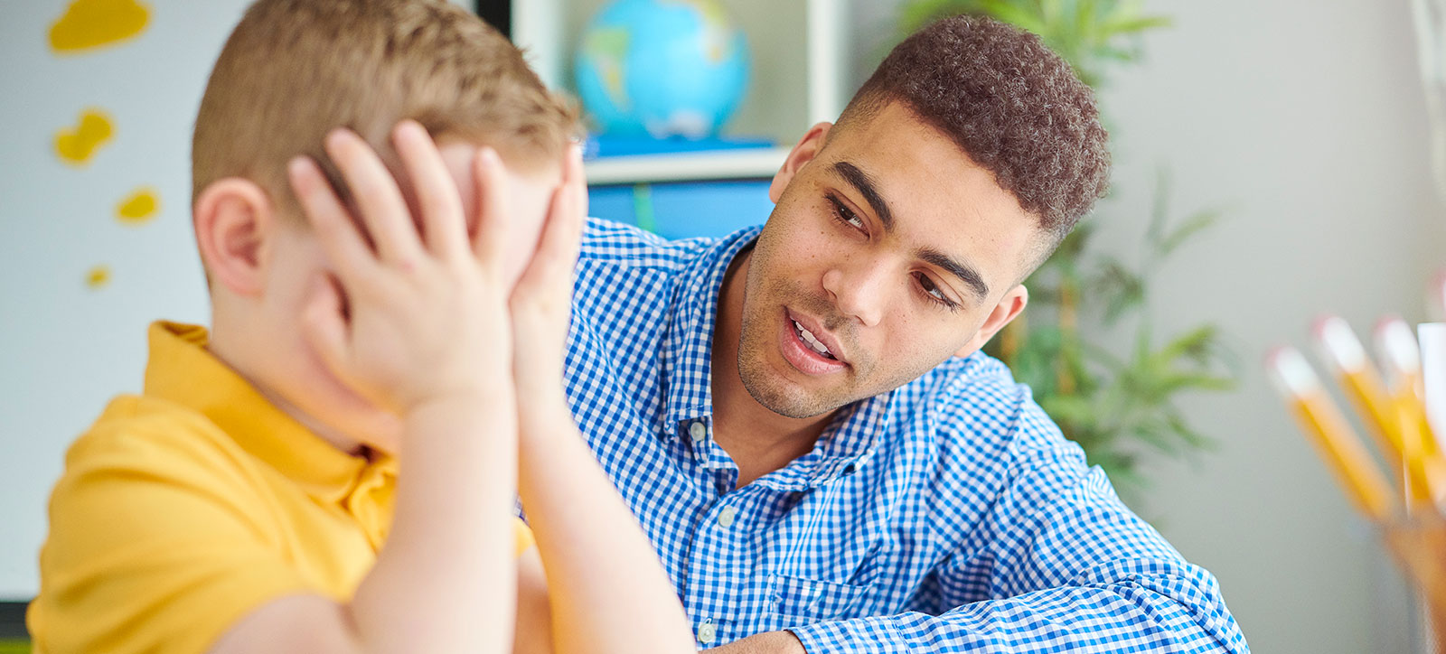 A young male teacher reassures a primary school boy who is sat with his head in his hands, looking distressed.