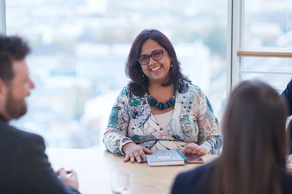 A person in business attire sits at a table smiling, with a notebook in front of them. Other individuals are partially visible in the foreground.